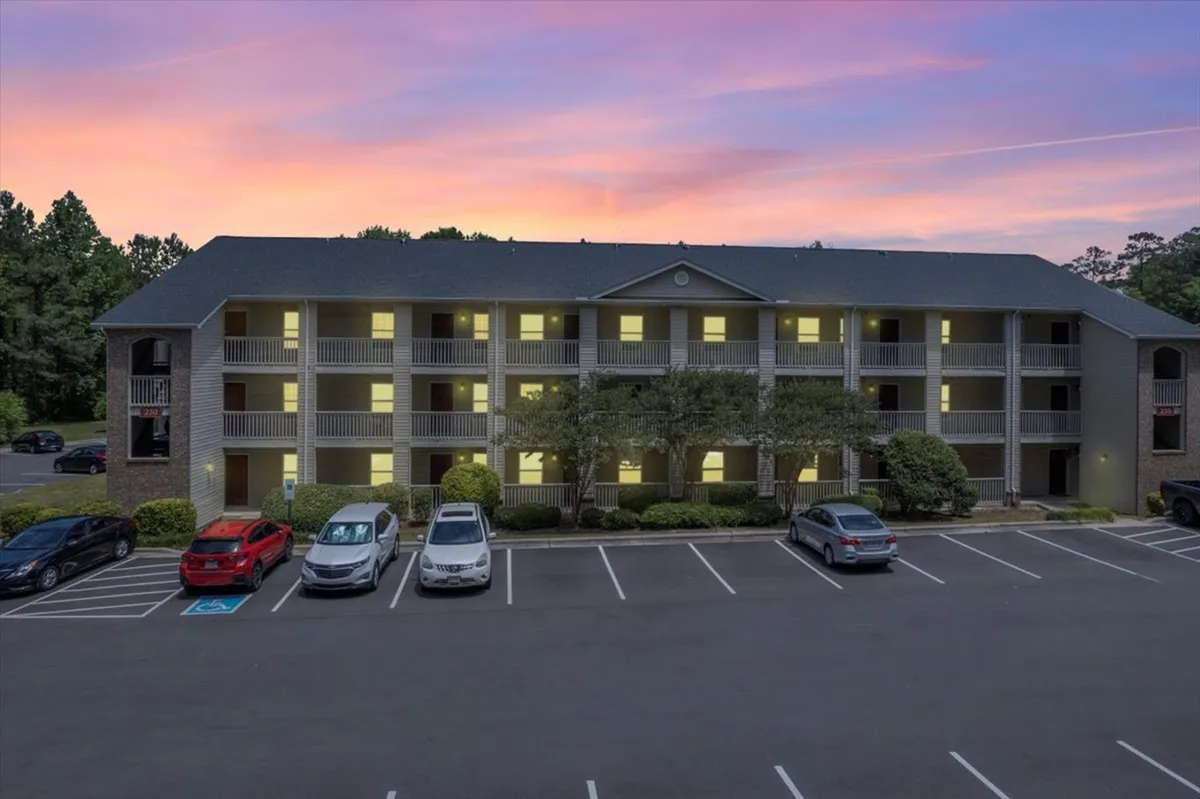 The image showcases the exterior of a modern apartment building in Fayetteville, North Carolina, with a spacious parking lot in the foreground and a vibrant sunset sky setting a warm, inviting mood. The balconies hint at opportunities for enjoying the evening outdoors, while the well-lit, clean surroundings suggest a welcoming community perfect for relaxation or socializing.