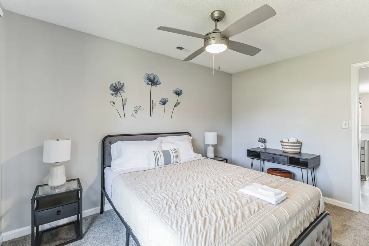 A tranquil bedroom featuring a plush bed adorned with decorative bedding, flanked by nightstands with lamps and thoughtful wall art featuring flowers. Soft natural light filters in, highlighting the calming and restful ambiance ideal for relaxation or a good night’s sleep.