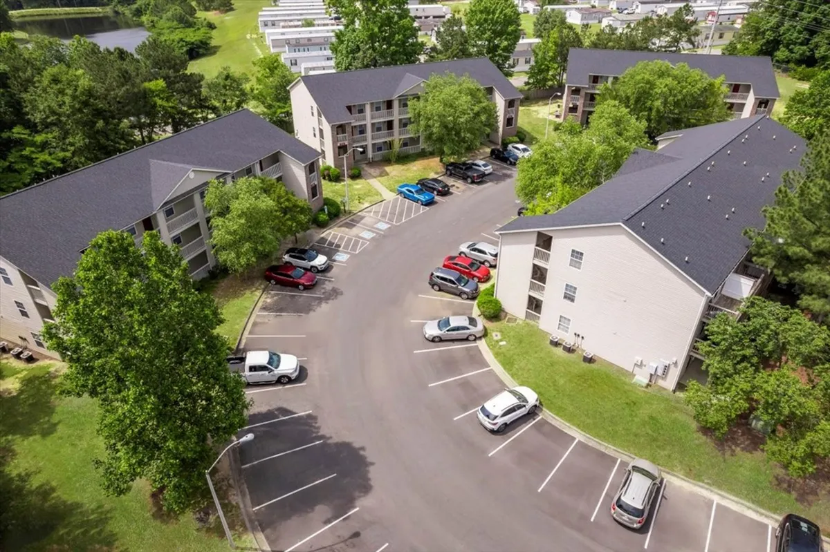 A view of apartment buildings nestled among green trees, with a visible parking area. The scene is bright and open, suggesting a safe, community-oriented atmosphere with convenient vehicle access.