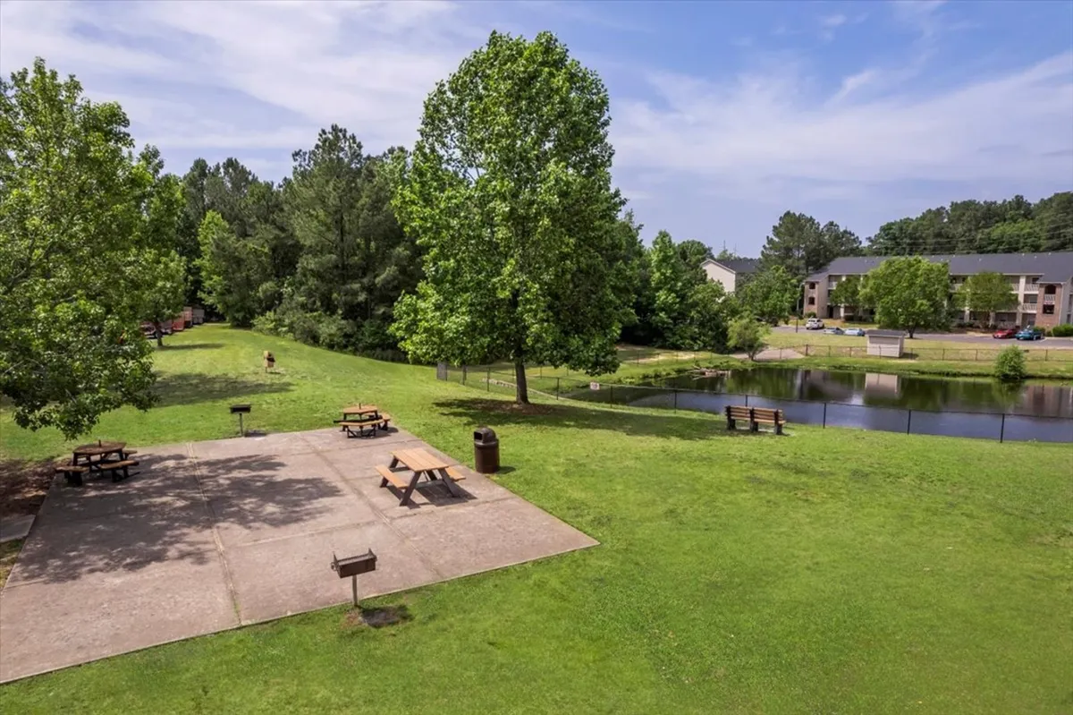 A tranquil exterior scene featuring a picnic area beside a pond, surrounded by mature trees and adjacent residential buildings. Residents can be seen enjoying the outdoors, engaging in activities like walking and picnicking.