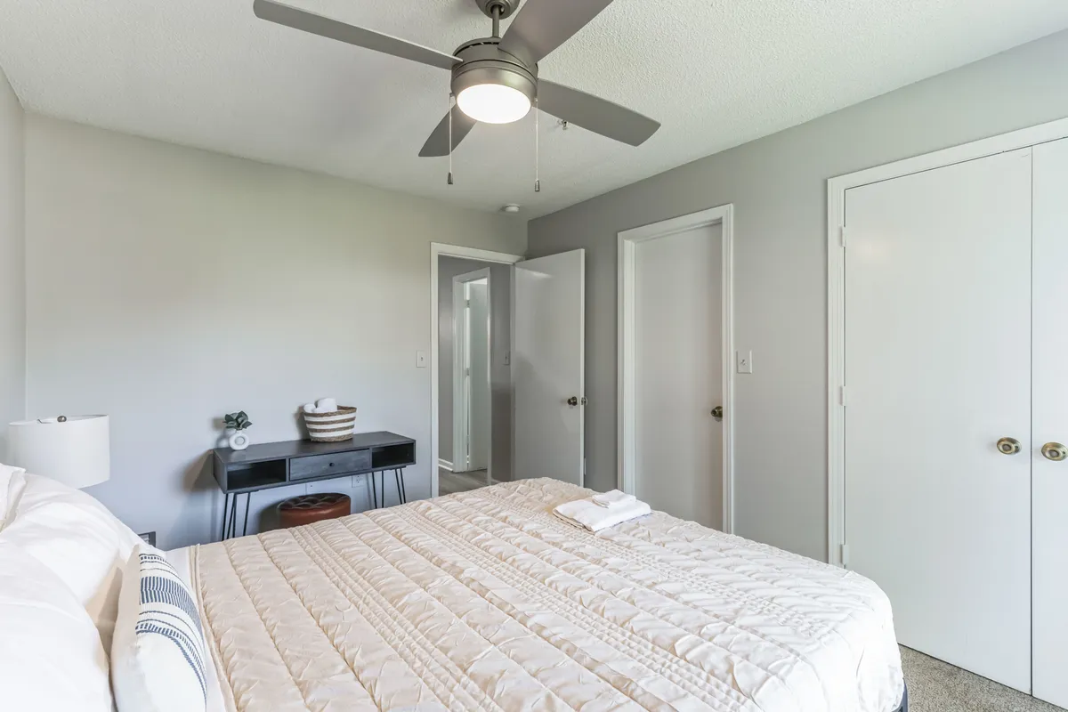 A serene bedroom featuring a bed with a white quilted cover, ceiling fan above, and a nightstand with a glowing lamp. The soft lighting and uncluttered design suggest a peaceful, home-away-from-home environment ideal for relaxation and rejuvenation.