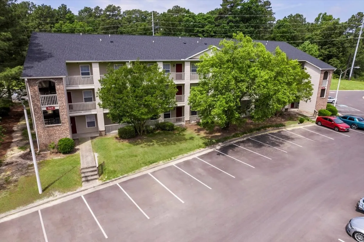 A welcoming exterior view of the apartment community, featuring lush green trees, balconies, and a well-maintained parking lot. The scene suggests a friendly environment and highlights the building’s inviting facade.