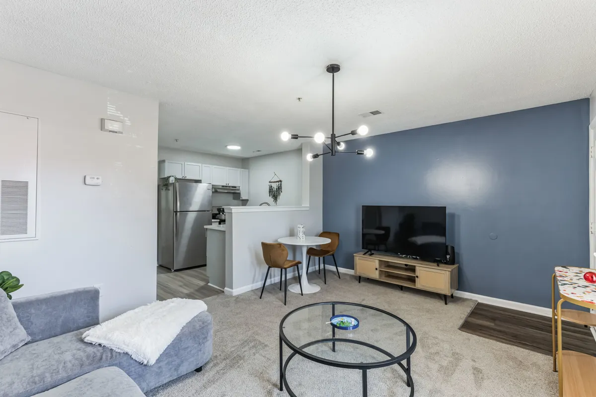 This image displays a welcoming living room featuring a plush sofa with a white throw blanket, a sleek glass coffee table, a TV stand with a television that invites you to stream your favorite shows, and a dining table with chairs off to the side. The space is modern yet cozy, with soft lighting and a layout designed for both relaxation and socializing.